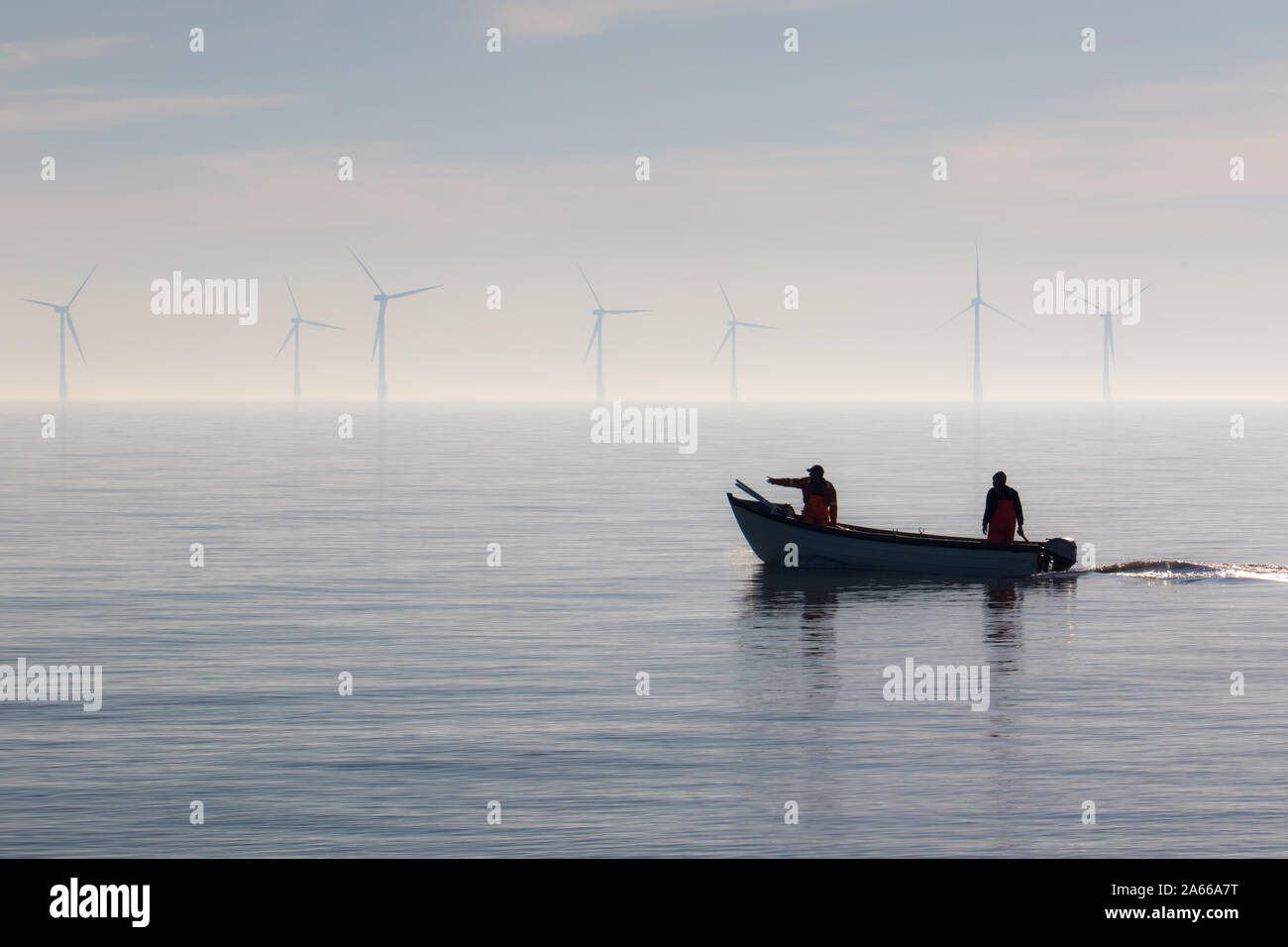 Sustainable resources alternative lifestyle. Small fishing boat with offshore wind turbines. Calm misty morning. Two fishermen heading out to sea. Tra Stock Photo