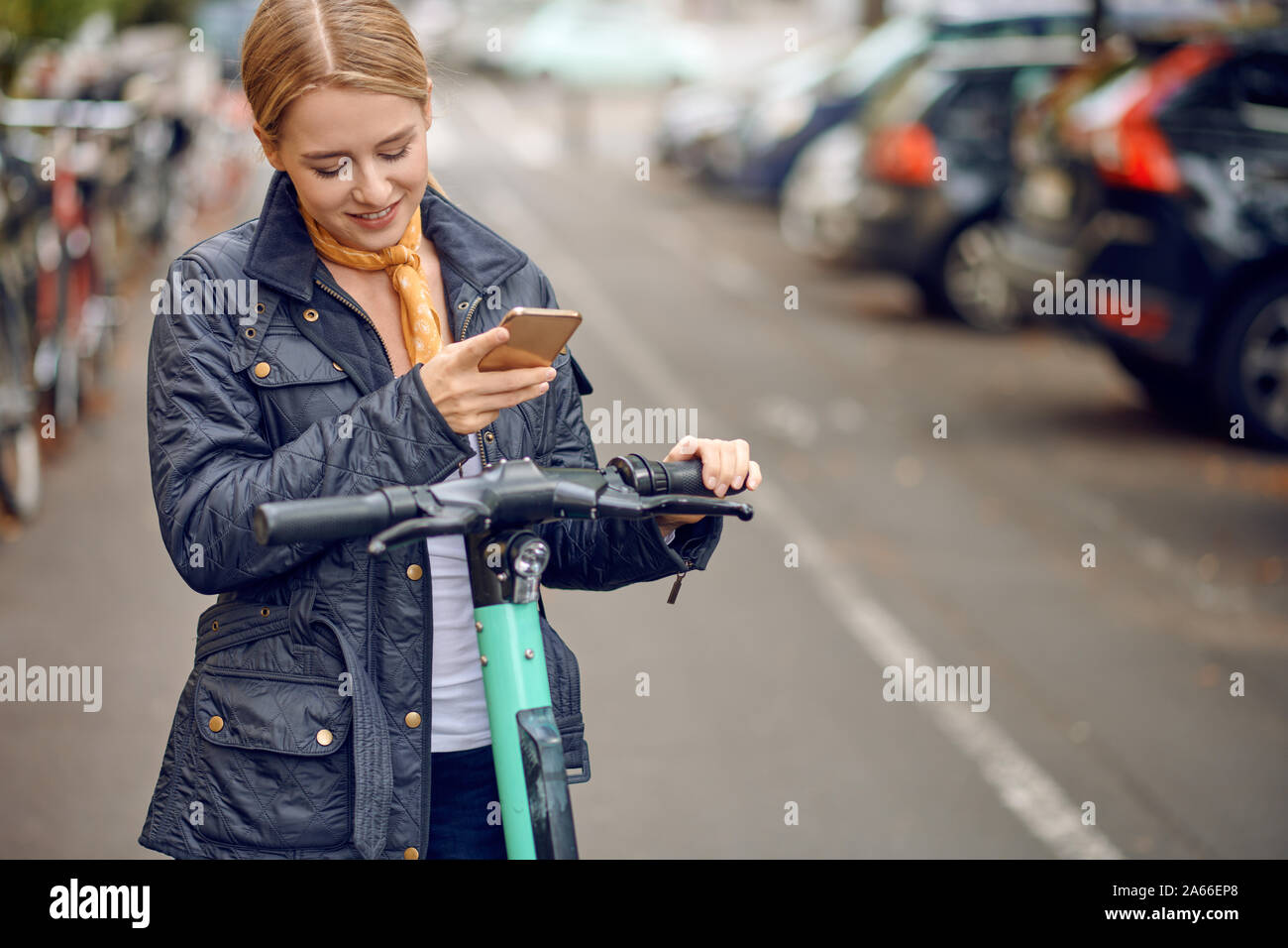 Happy young woman in casual grey jacket looking at camera and smiling, standing in the street with an e-scooter with mobile phone in her hands. Stock Photo