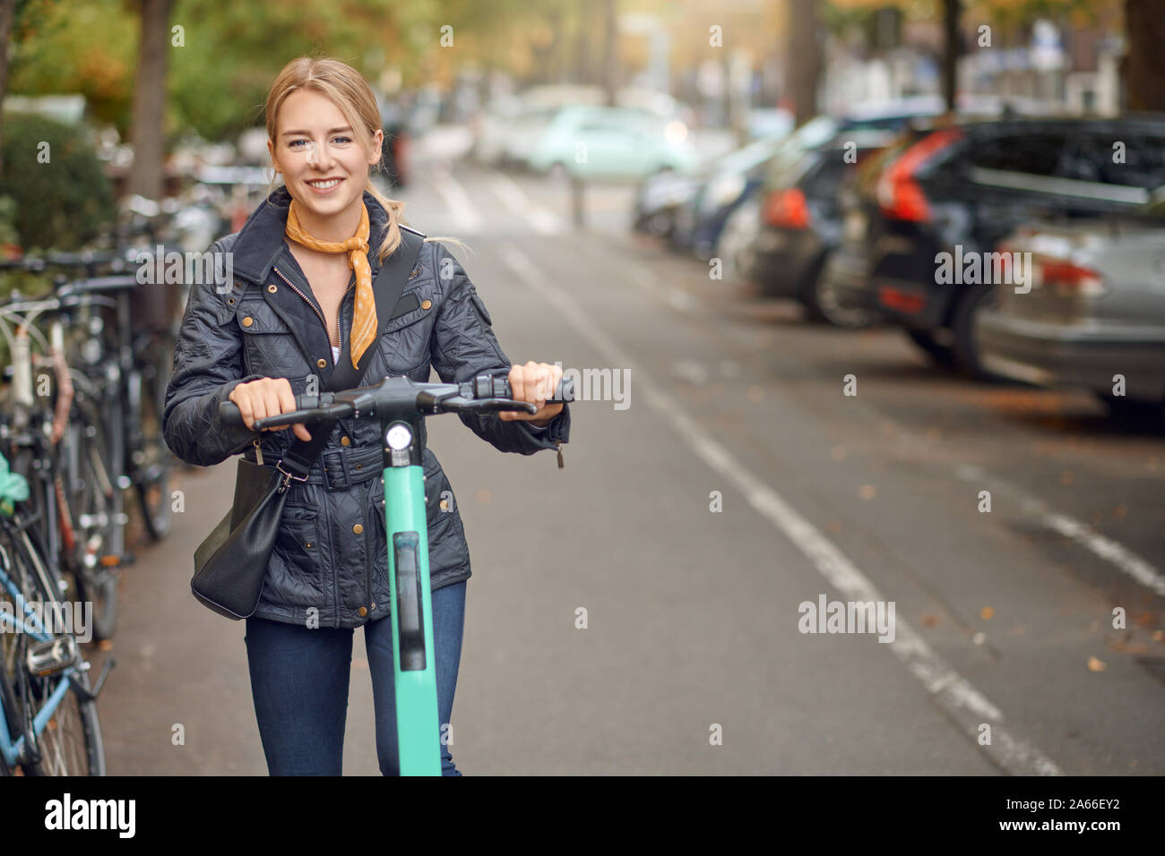 Young happy blond woman riding an electric scooter in the city, smiling at the camera, in autumn Stock Photo
