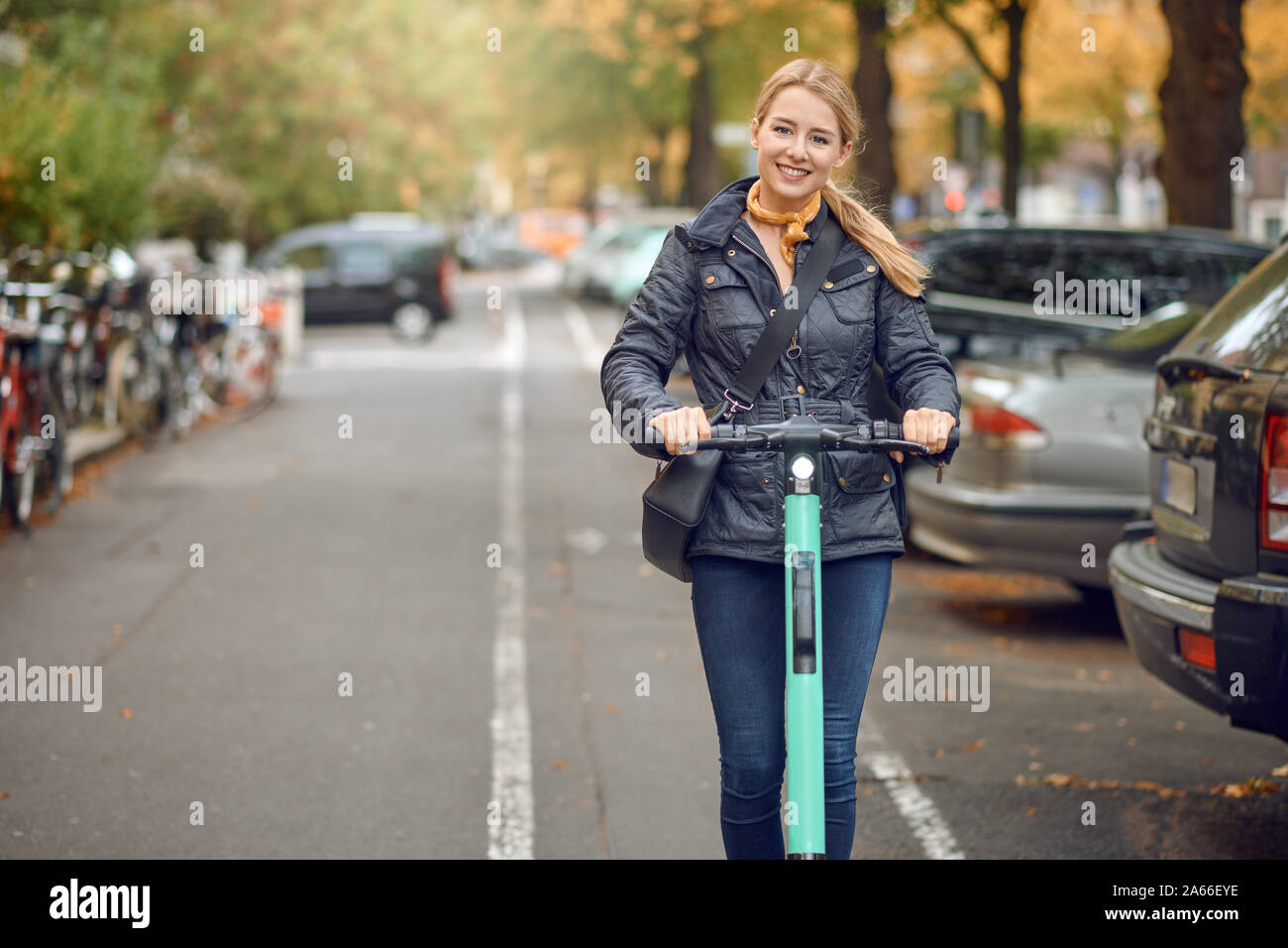 Young happy blond woman riding an electric scooter in the city, smiling at the camera, in autumn Stock Photo