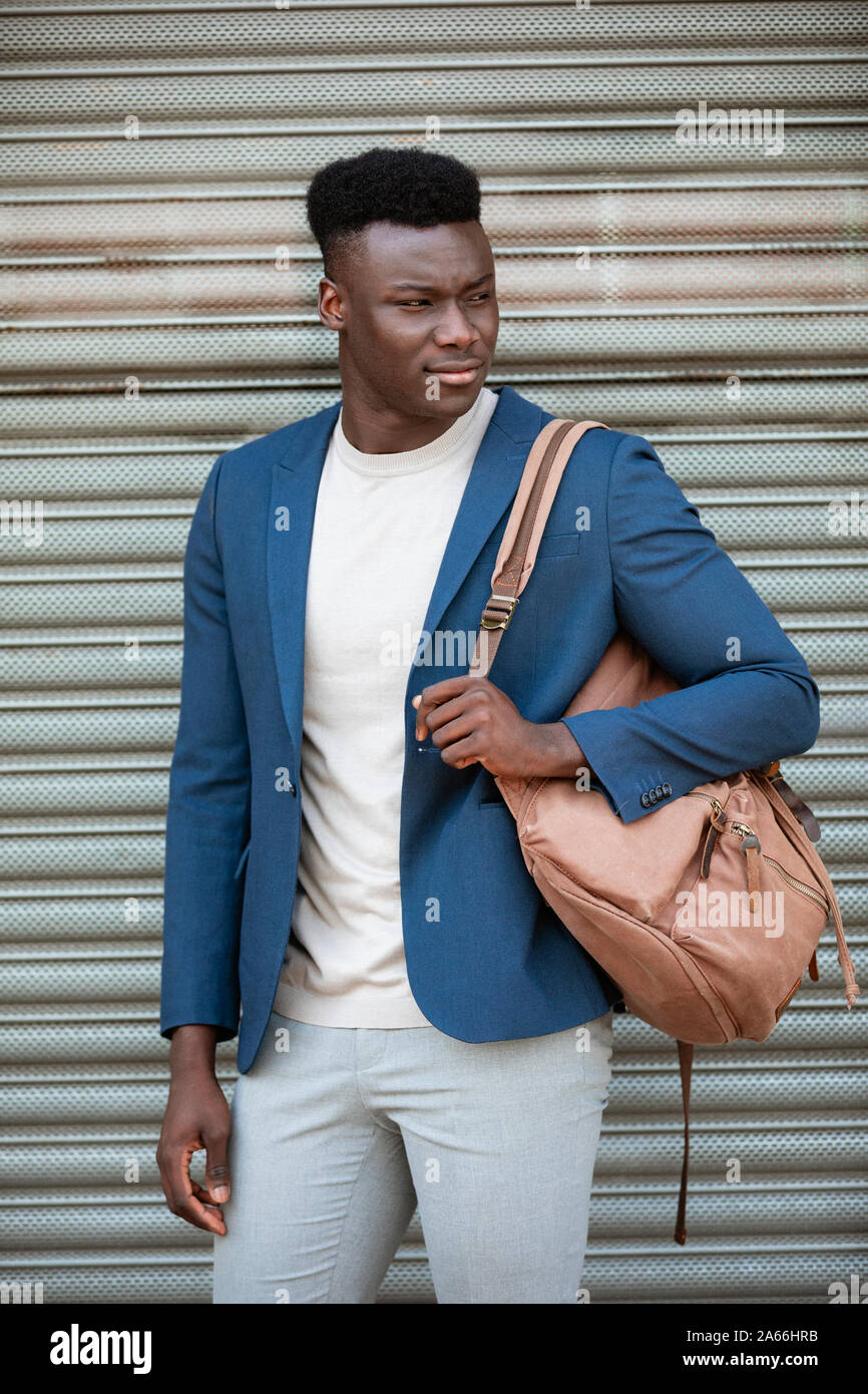 Three quarter length shot of a businessman standing in front of a shutter in the street. He is wearing smart casual clothing while holding a satchel o Stock Photo