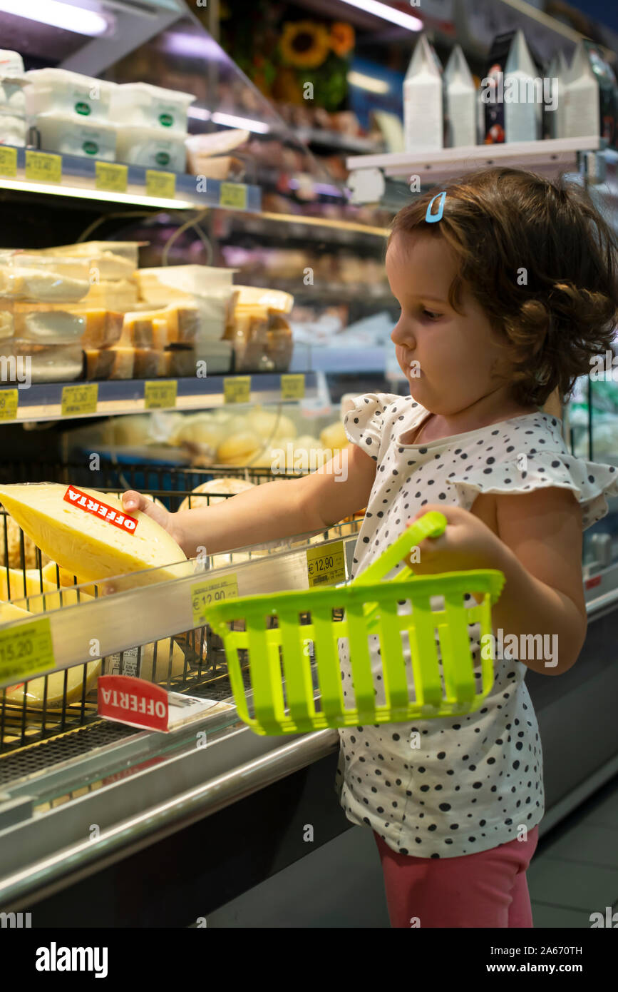 Little girl buying cheese in supermarket. Child hold small basket in supermarket and select cheese from store showcase. Concept for children selecting Stock Photo