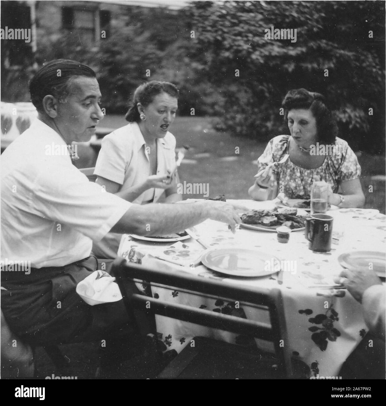 Three members of a Jewish-American family sit outdoors around a small table and eat a picnic, with two women in mid conversation and man reaching for a plate of food, New York City, New York, 1948. () Stock Photo
