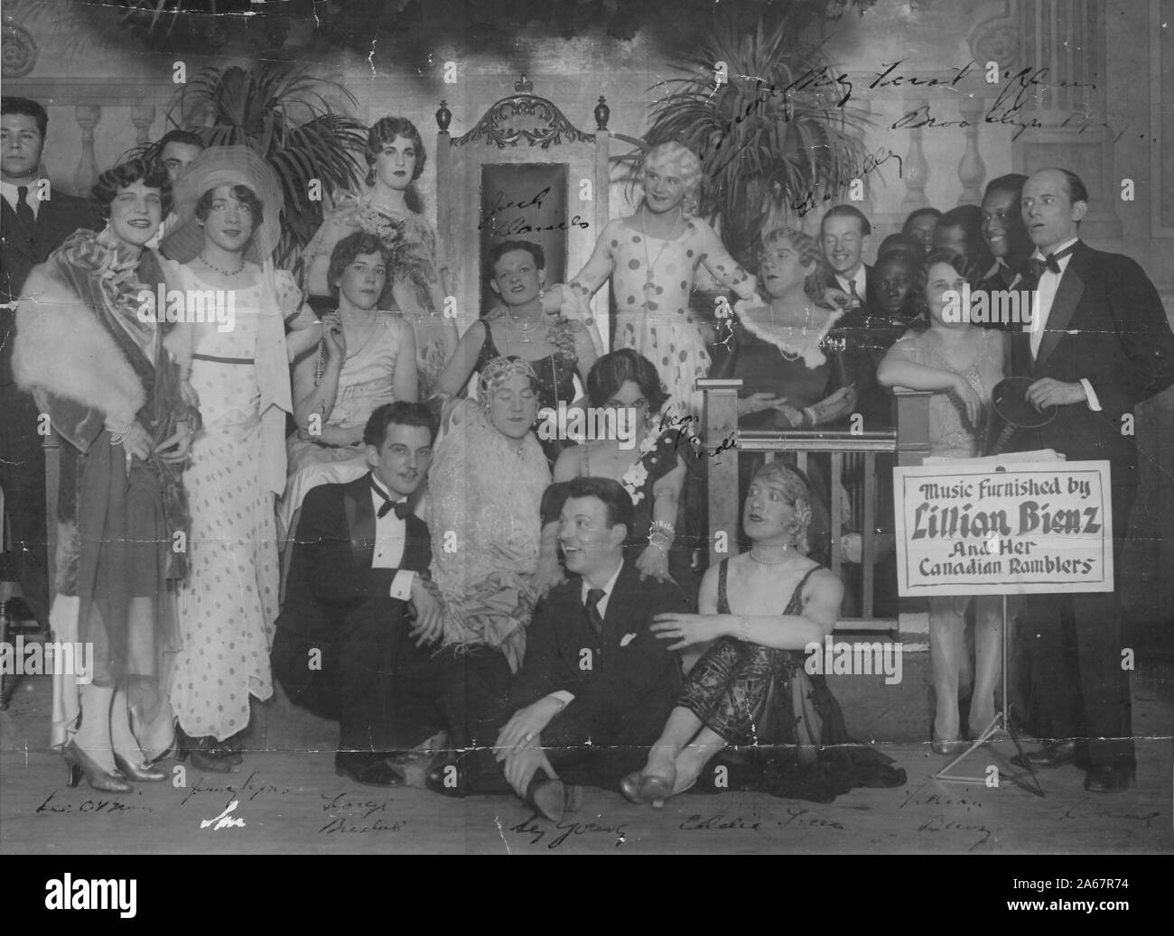 A group of drag queens, or men dressed in drag, pose for a group photo in front of a backdrop with a large crown prop chair, many in dresses and elegant clothing, with sign noting that music was provided by Lillian Benz and her Canadian Ramblers, a musical group, Brooklyn, New York, 1929. () Stock Photo