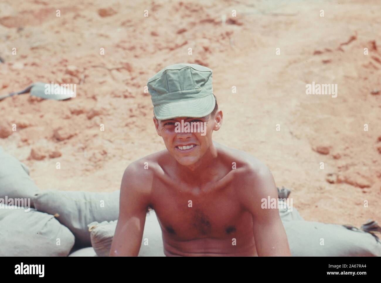 High-angle close-up of a shirtless American serviceman, from the chest up, sitting on sandbags and squinting up toward the camera, Vietnam, 1965. () Stock Photo