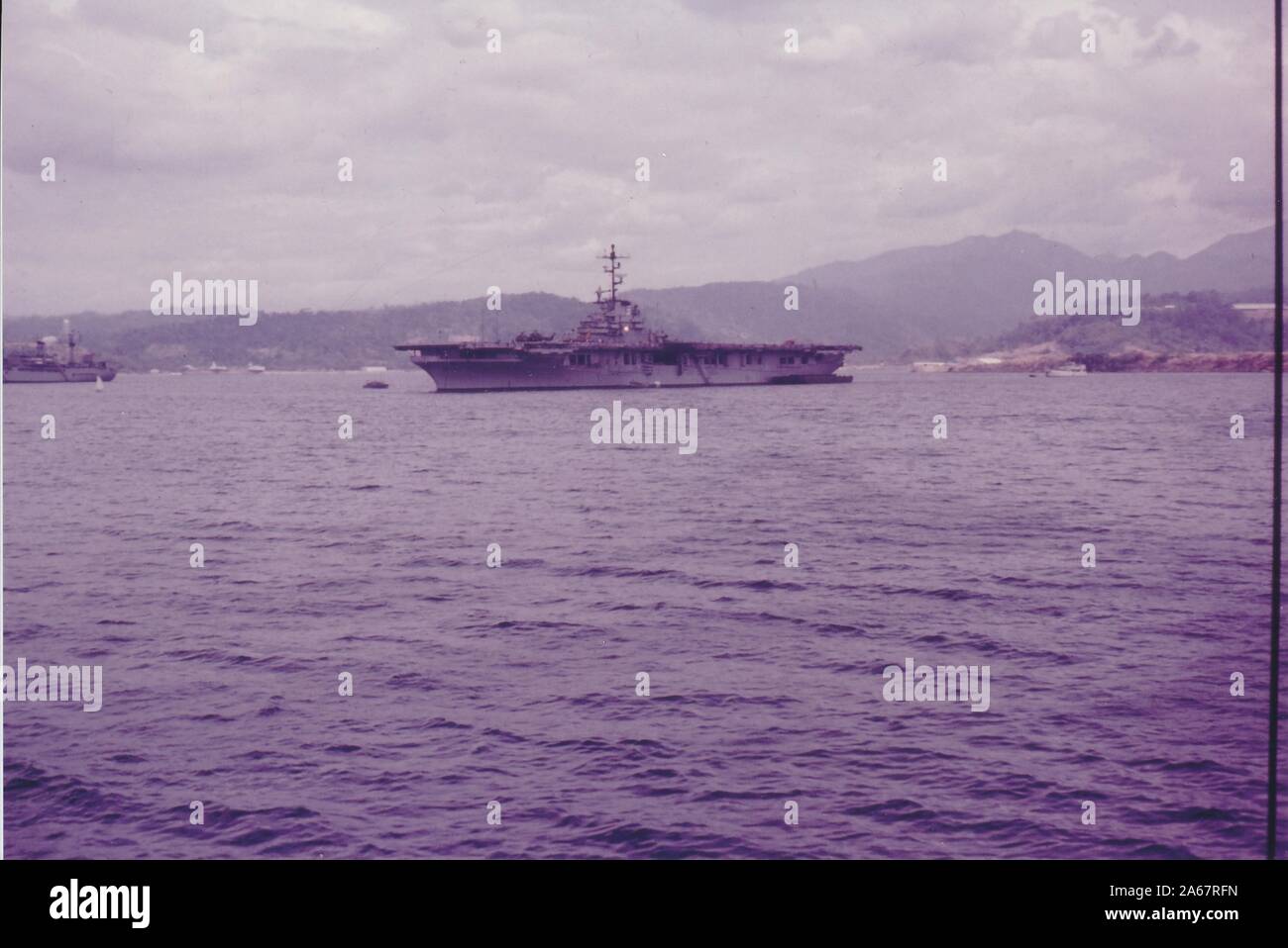 Wide shot, across open water, of an American military vessel near a mountainous coastline on an overcast day, photographed during the Vietnam War, 1965. () Stock Photo