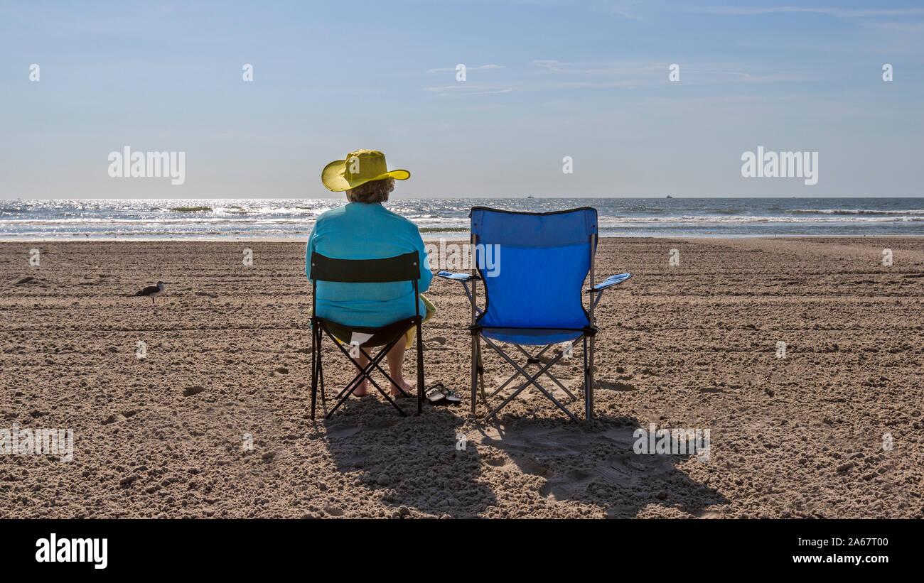 A senior woman enjoying sitting in a beach chair  in the sand on the beach by the ocean shore. Stock Photo