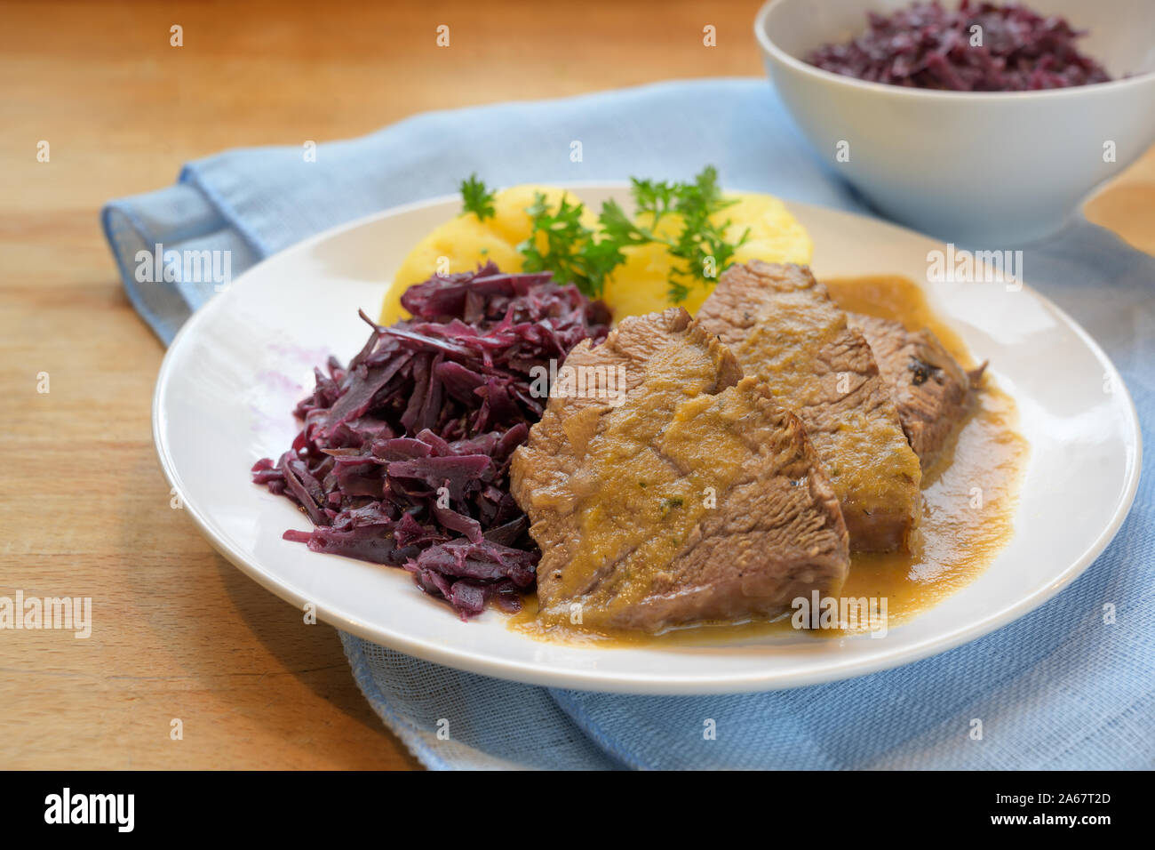 braised beef with potatoes, red cabbage and parsley garnish on a plate, blue napkin on a wooden table, selected focus, very narrow depth of field Stock Photo