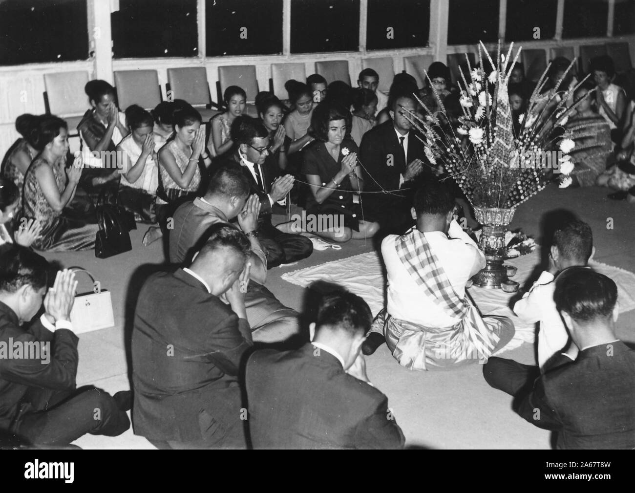 A group of formally dressed men and women sit on the floor, with their palms in the wai position, during a Baci ceremony at a joint function for the American military and the Royal Thai Army, photographed during the Vietnam War, 1968. () Stock Photo