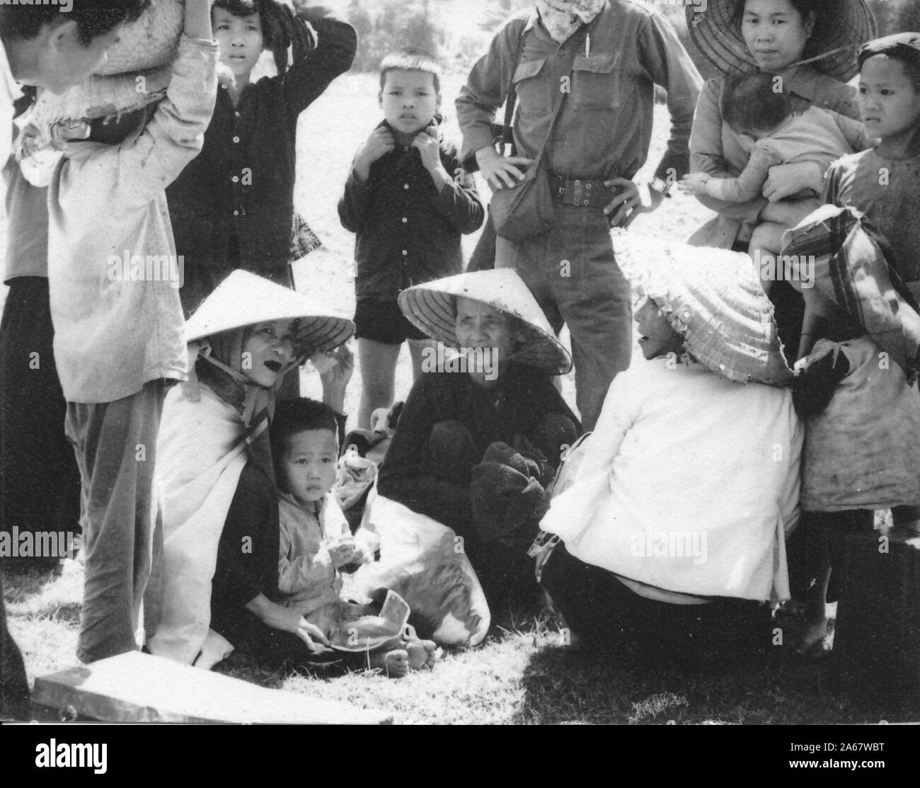 Close-up of a crowd of Vietnamese women and children, sitting and standing outside on a sunny day, flanking a standing, uniformed man whose head is off-camera, Vietnam, 1965. () Stock Photo