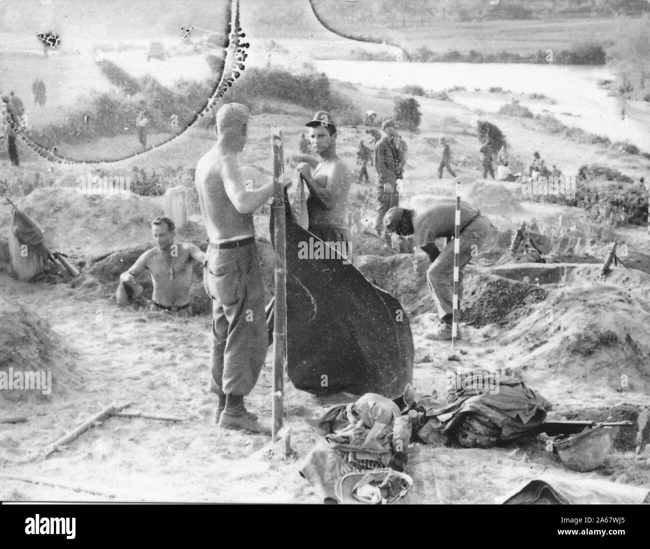 A group of uniformed and shirtless American soldiers dig trenches and organize other fortifications, while preparing a camp on an open hillside on a sunny day, Vietnam, 1965. () Stock Photo