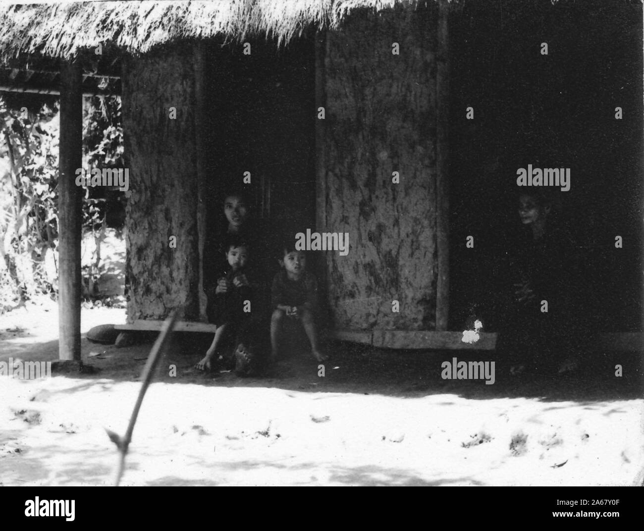 A Vietnamese woman and two small children sit in the shady doorway of a thatch-roofed building on a sunny day, Vietnam, 1965. () Stock Photo