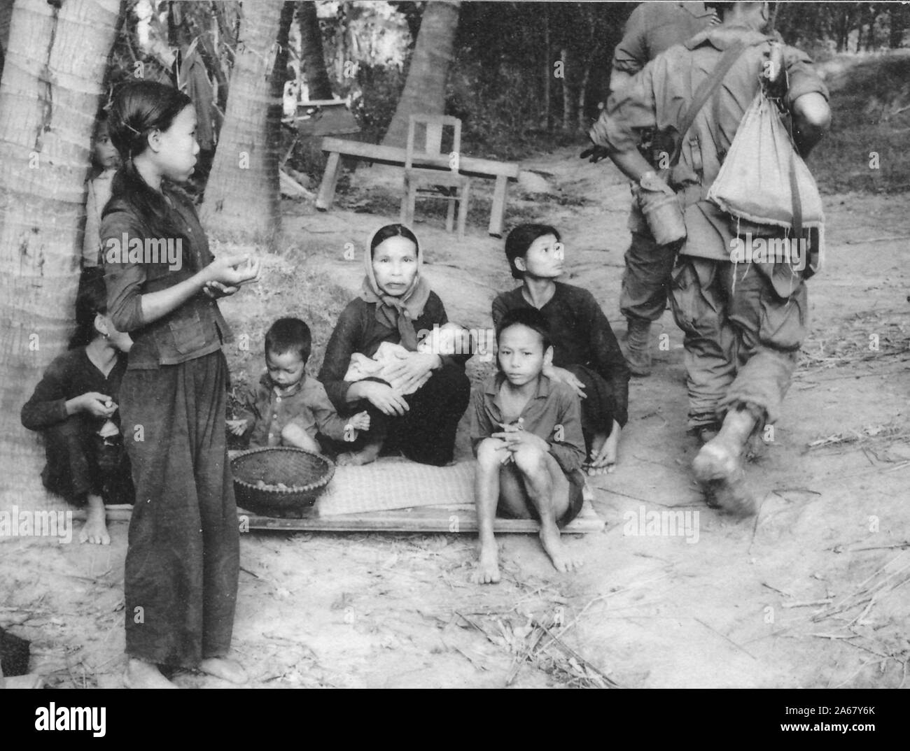 A small group of Vietnamese women and children sit outside on a wooden plinth in a shady area, some face the camera while others watch uniformed soldiers walk past at right, Vietnam, 1965. () Stock Photo