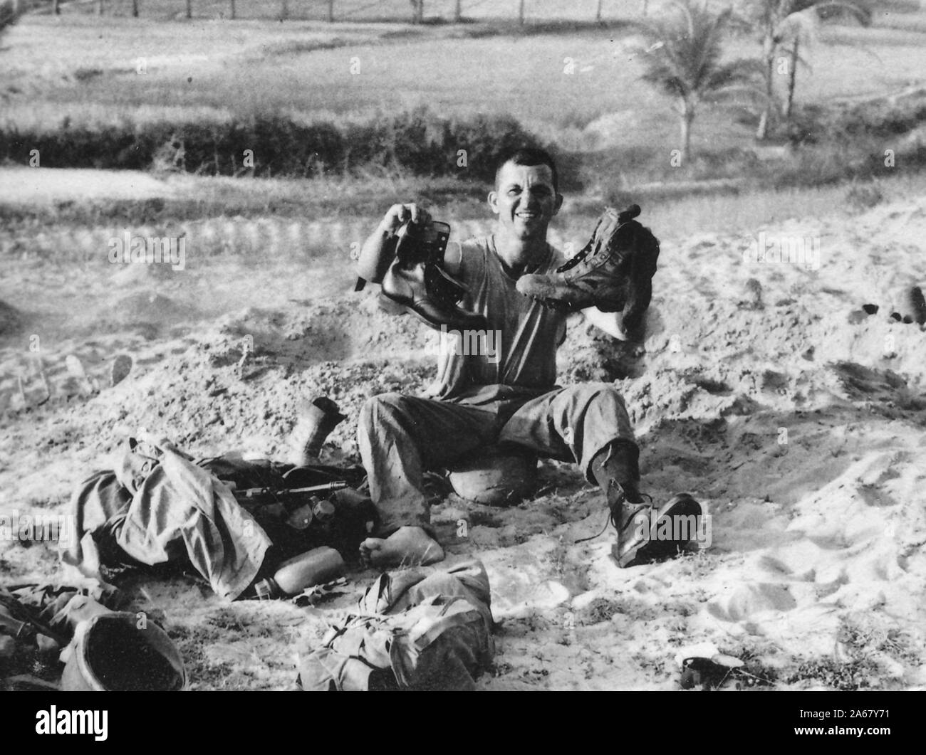 An American serviceman sits outside on a sandy hillside on a sunny day, making a face at the camera, while holding up a new boot in one hand and an old boot in the other, Vietnam, 1965. () Stock Photo