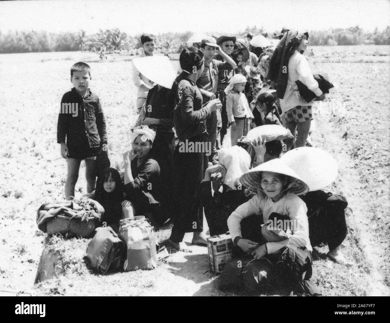 A small crowd of Vietnamese women and children, seated and standing in an open field on a sunny day, with a pair of soldiers standing in between them, Vietnam, 1965. () Stock Photo