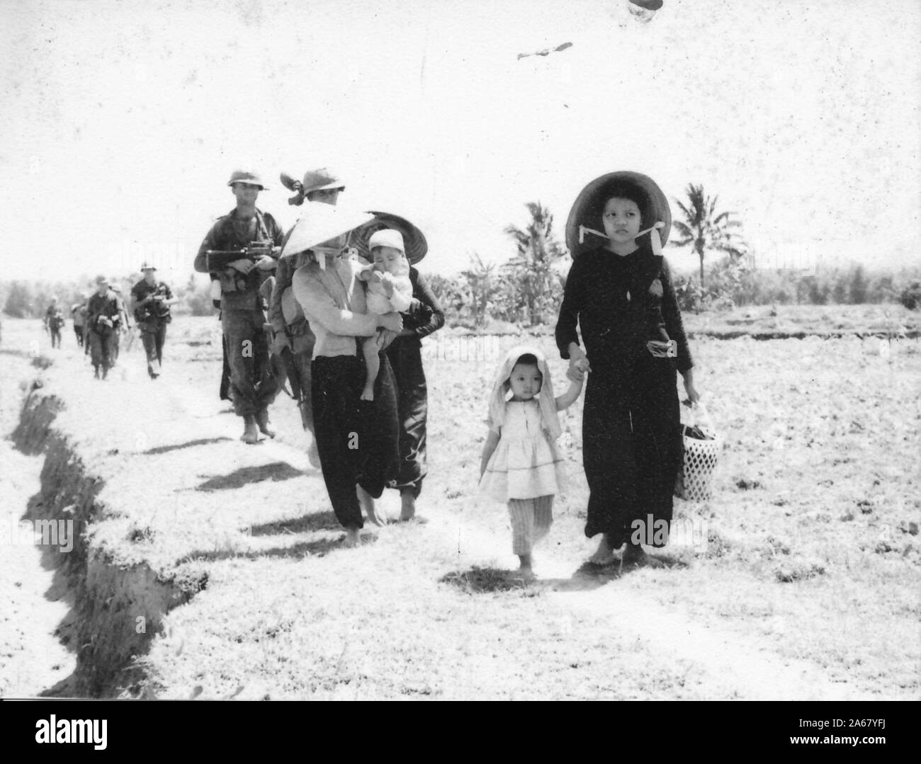 Civilian Vietnamese women and children, walk on a dirt path on a sunny day, in front of a staggered line of American servicemen, Vietnam, 1965. () Stock Photo