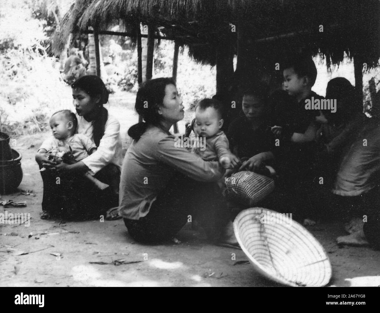 Civilian Vietnamese women and children sit together in the shade of thatched-roof hut on a sunny day, Vietnam, 1965. () Stock Photo