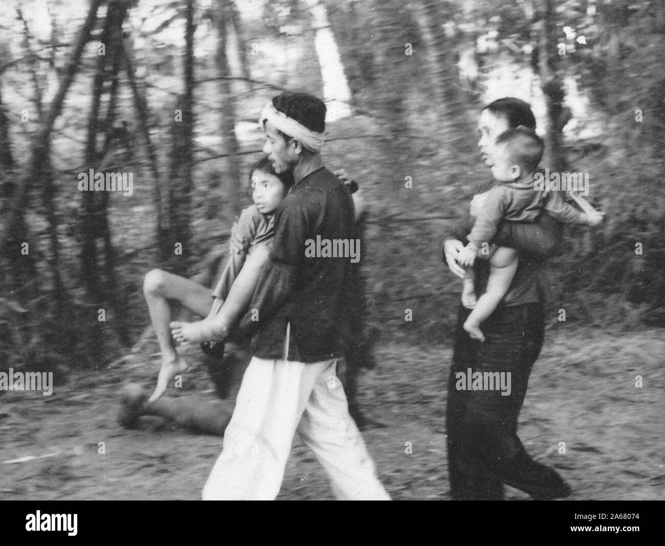 Profile shot, in a forested area, of a Vietnamese man carrying a young girl and a woman carrying a baby, Vietnam, 1965. () Stock Photo