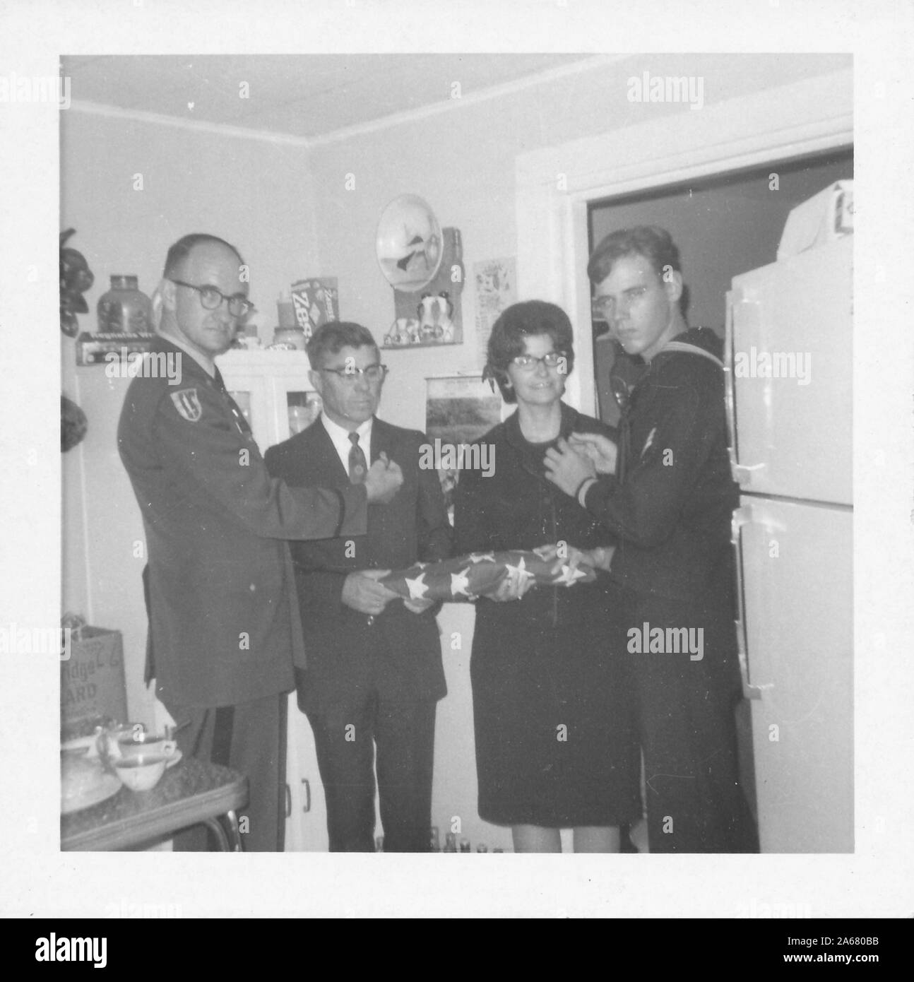 A civilian man and woman, likely the parents of a deceased soldier, stand in a small kitchen holding a folded American flag while two uniformed American servicemen hold medals near their chests, United States of America, 1965. () Stock Photo