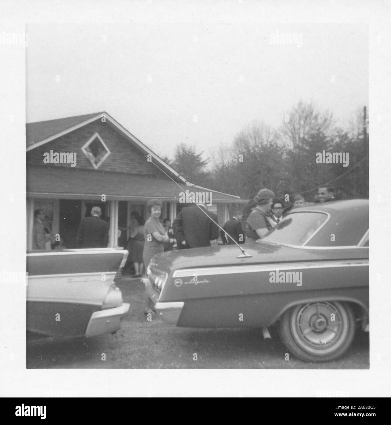 A small group of people stand outside near a brick building on an overcast day, with the tailfins of a Ford Thunderbird and the trunk of a Chevy Impala in the foreground, likely following a military funeral, United States of America, 1965. () Stock Photo