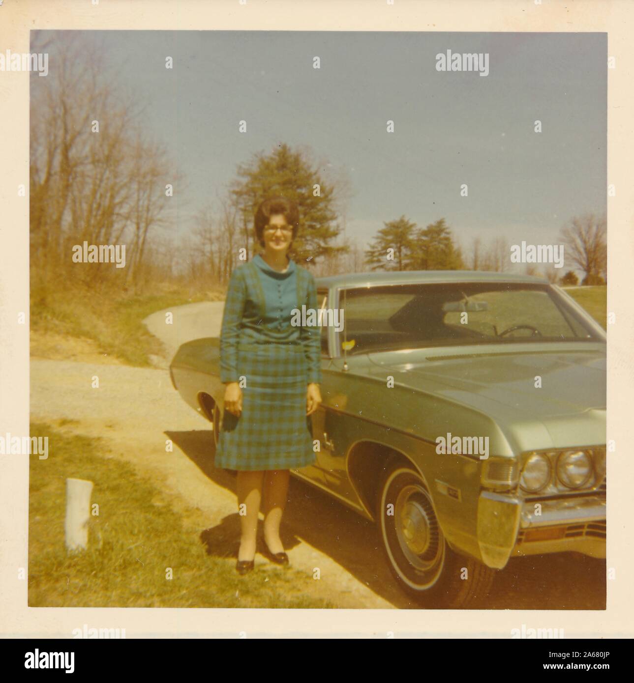 Full-length shot of a woman, wearing a vintage checked skirt suit and glasses, posing next to a Chevrolet Impala parked on a deserted country road on a sunny day, United States of America, 1965. () Stock Photo