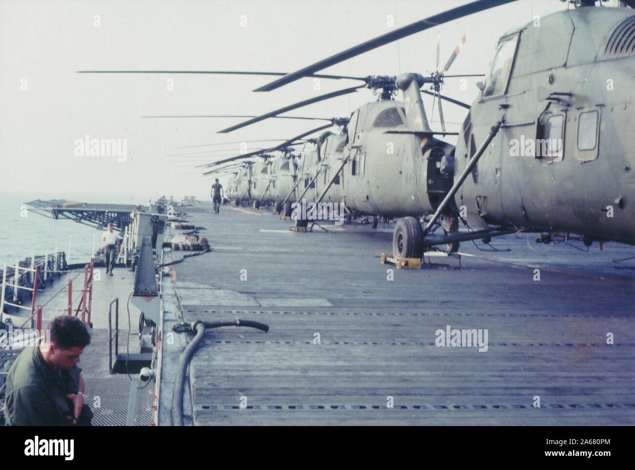 Two American servicemen stand and walk near a row of helicopters docked on the deck of a military aircraft carrier located near the coast of Vietnam, 1965. () Stock Photo