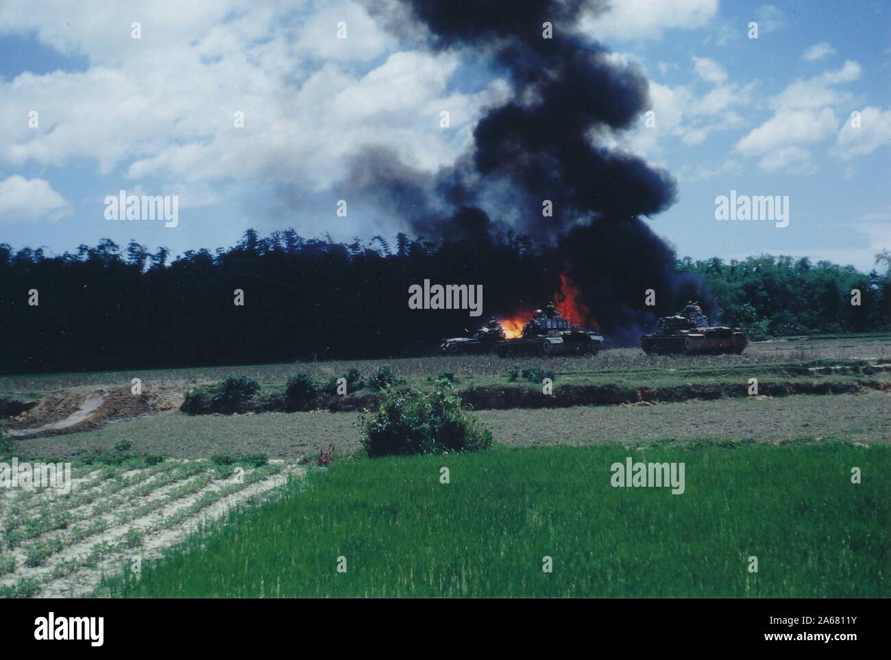 Slightly angled wide shot, across open fields, of military vehicles near a flaming and smoking explosion at the edge of a forested area, Vietnam, 1965. () Stock Photo