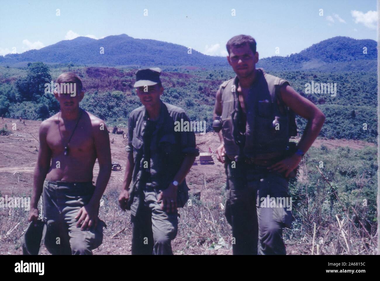 Three-quarter length shot of three American servicemen, facing the camera, while standing outside on a sunny day on the rise of a small hill with a cleared camp in the background, Vietnam, 1965. () Stock Photo