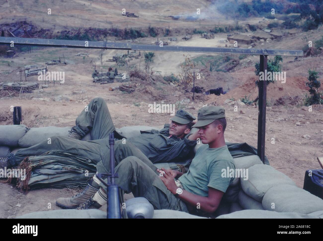 Profile shot of two American servicemen lounging in a sandbag bunker, with more bunkers, tanks, and camp tents in the background, Vietnam, 1965. () Stock Photo