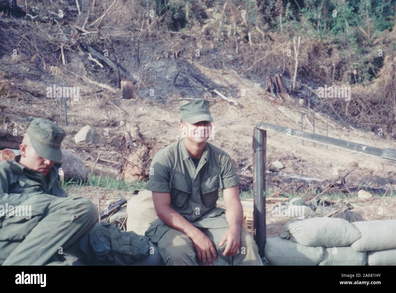 Two American servicemen outside on a sunny day, one reclining and looking down, the other sitting on sandbags and smiling, with charred underbrush in the background, Vietnam, 1965. () Stock Photo
