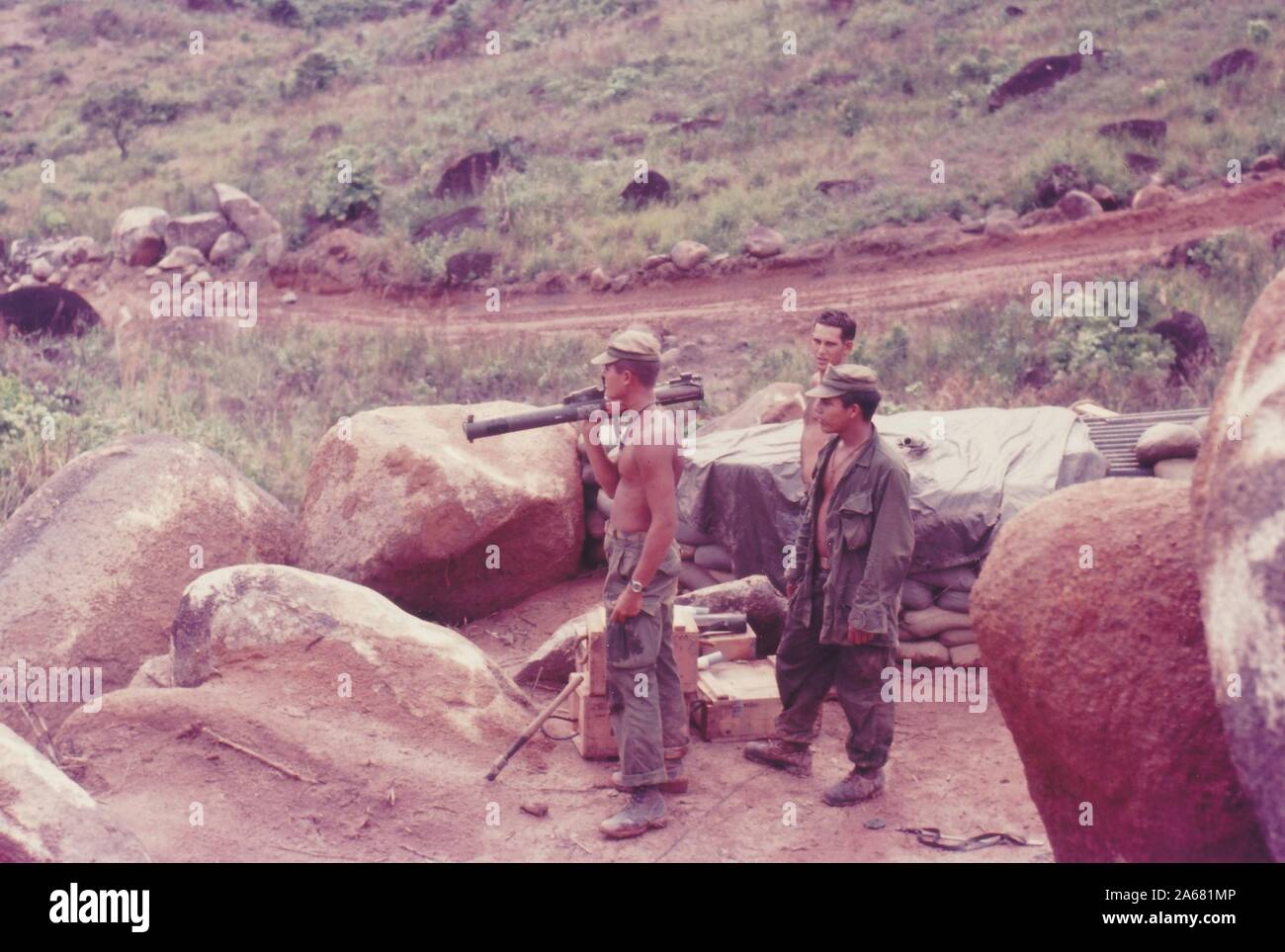 High-angle profile shot of three American servicemen, one with a bazooka balanced on his shoulder, standing outside in a rural area near boulders and sandbags, Vietnam, 1965. () Stock Photo