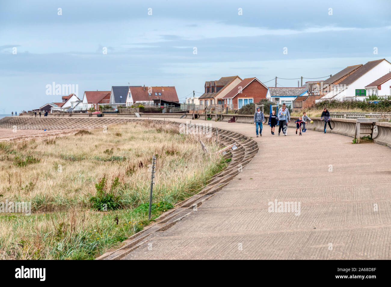 People walking along the protective sea wall built to avoid flooding in front of seaside bungalows facing The Wash at Heacham in West Norfolk. Stock Photo