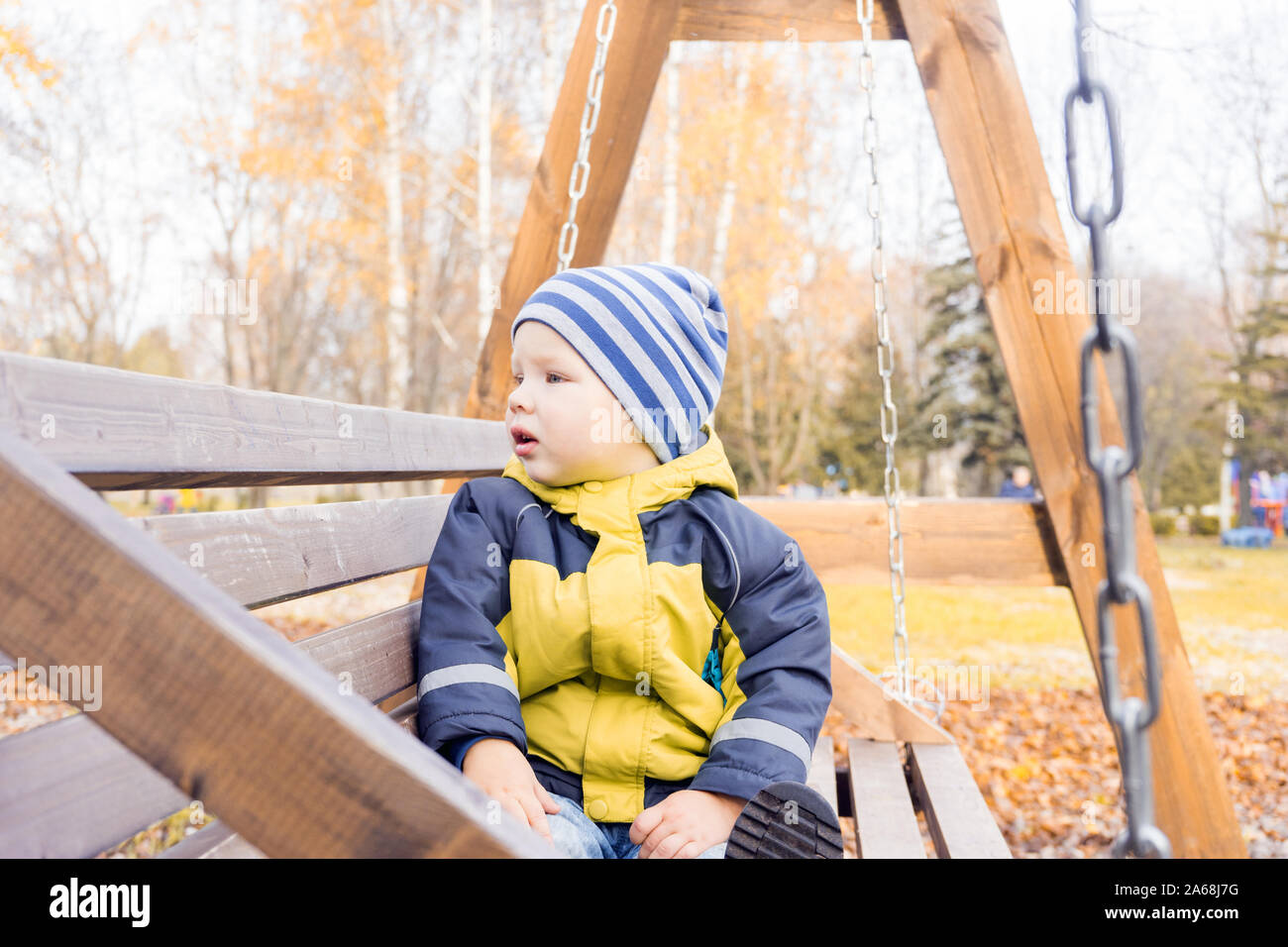 toddler child looks around with interest swinging on wooden swing on Sunny autumn day in city Park. Stock Photo