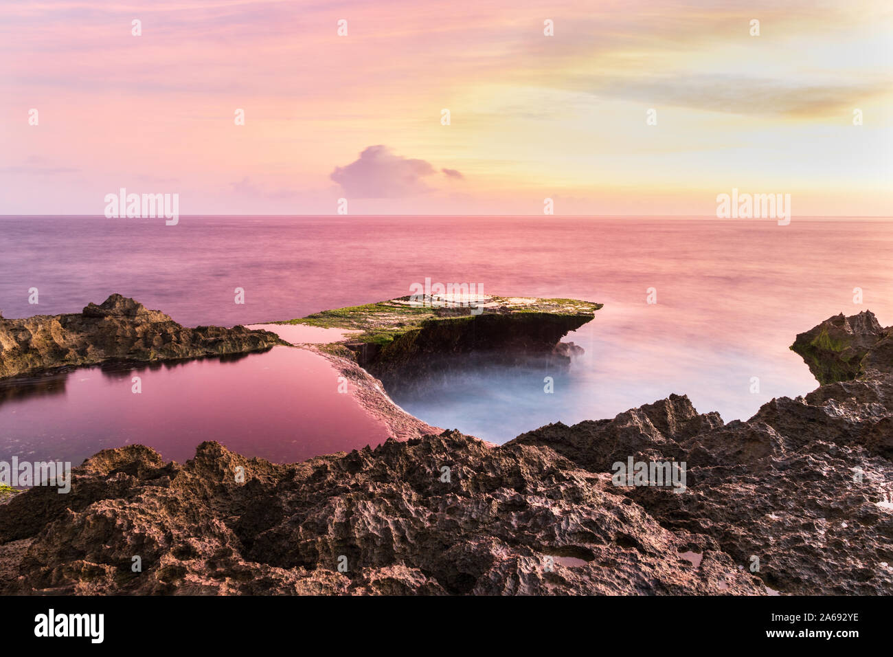 Devil's Tear at sunset, island of Nusa Lembongan, Bali, Indonesia. Rocky shore in foreground. Tidepool and ocean pink from setting sun's reflection. Stock Photo