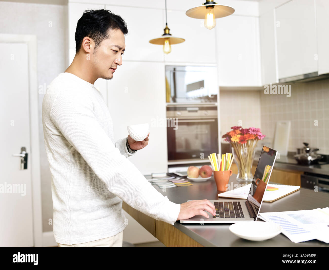 young asian man working from home standing by kitchen counter using laptop computer Stock Photo