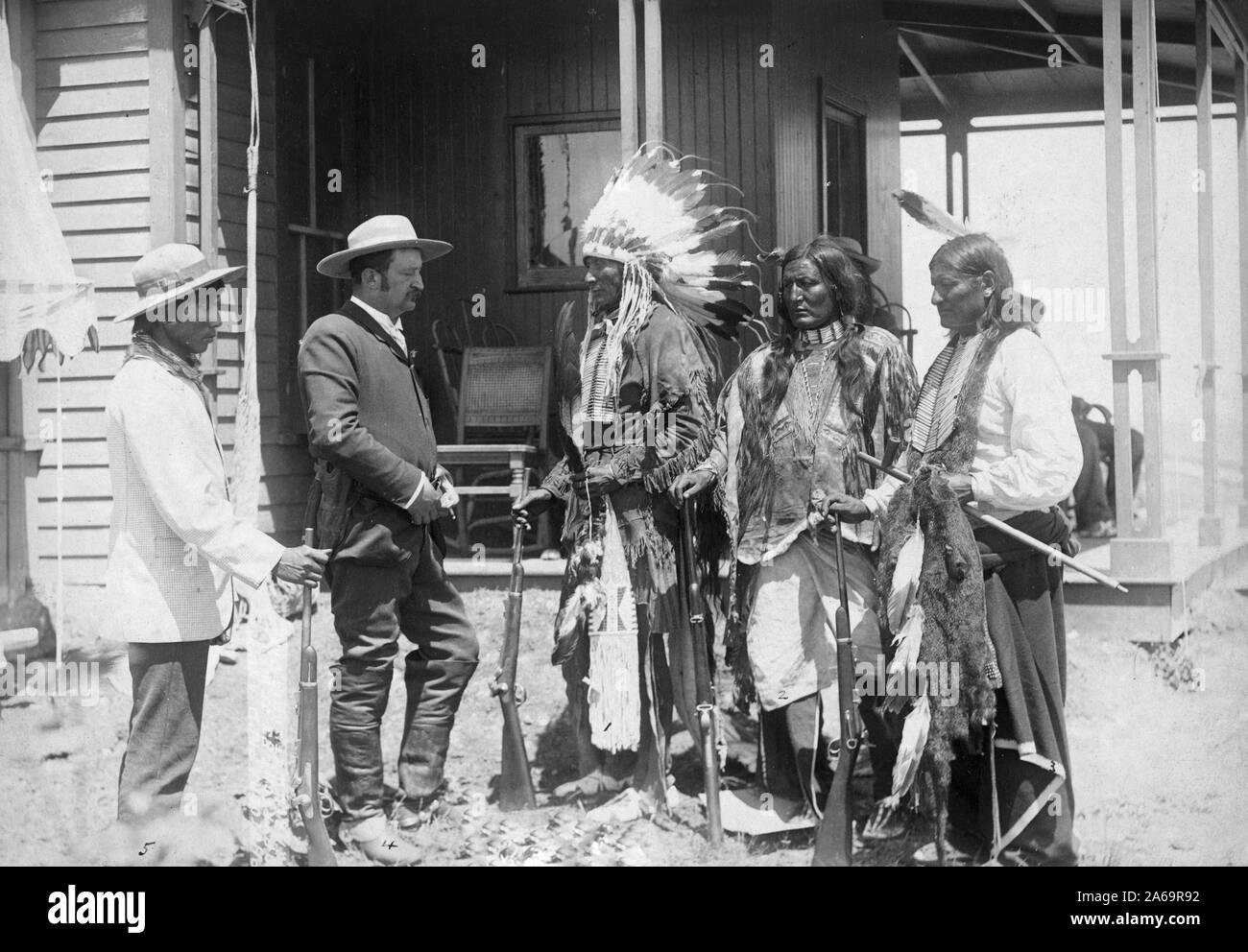 Three Cheyenne men wearing ceremonial clothing and holding rifles, greeting a white man in a suit and his interpreter in front of a building. Stock Photo