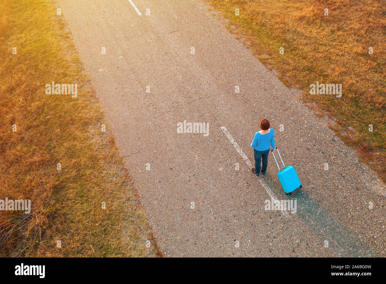 Woman pulling travel suitcase luggage on road in autumn sunset, high angle view from drone pov Stock Photo