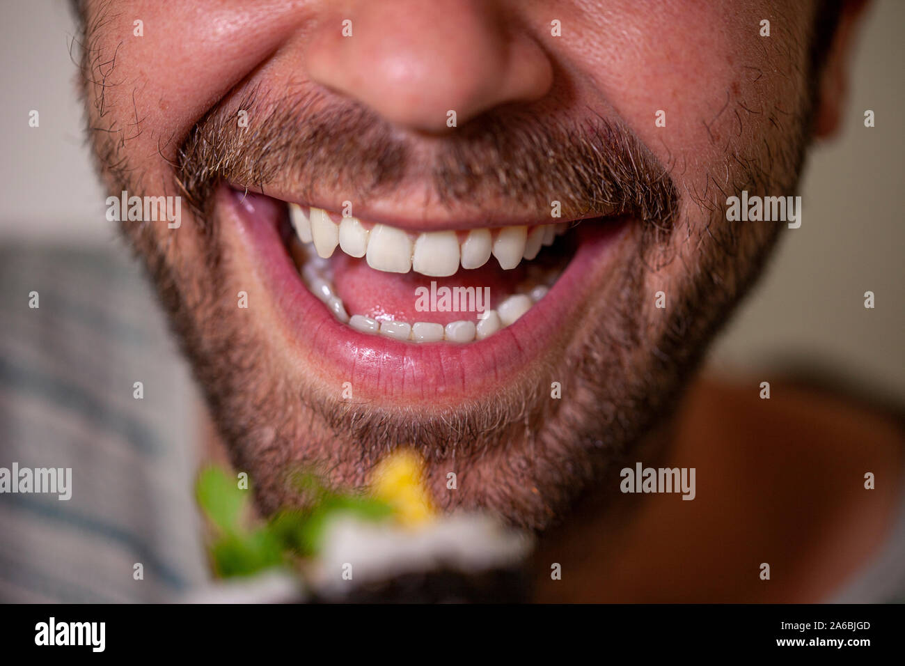 Close up of a smiling mouth of a bearded caucasian man about to eat a homemade sashimi sushi roll with chopsticks which is out of focus in the front Stock Photo