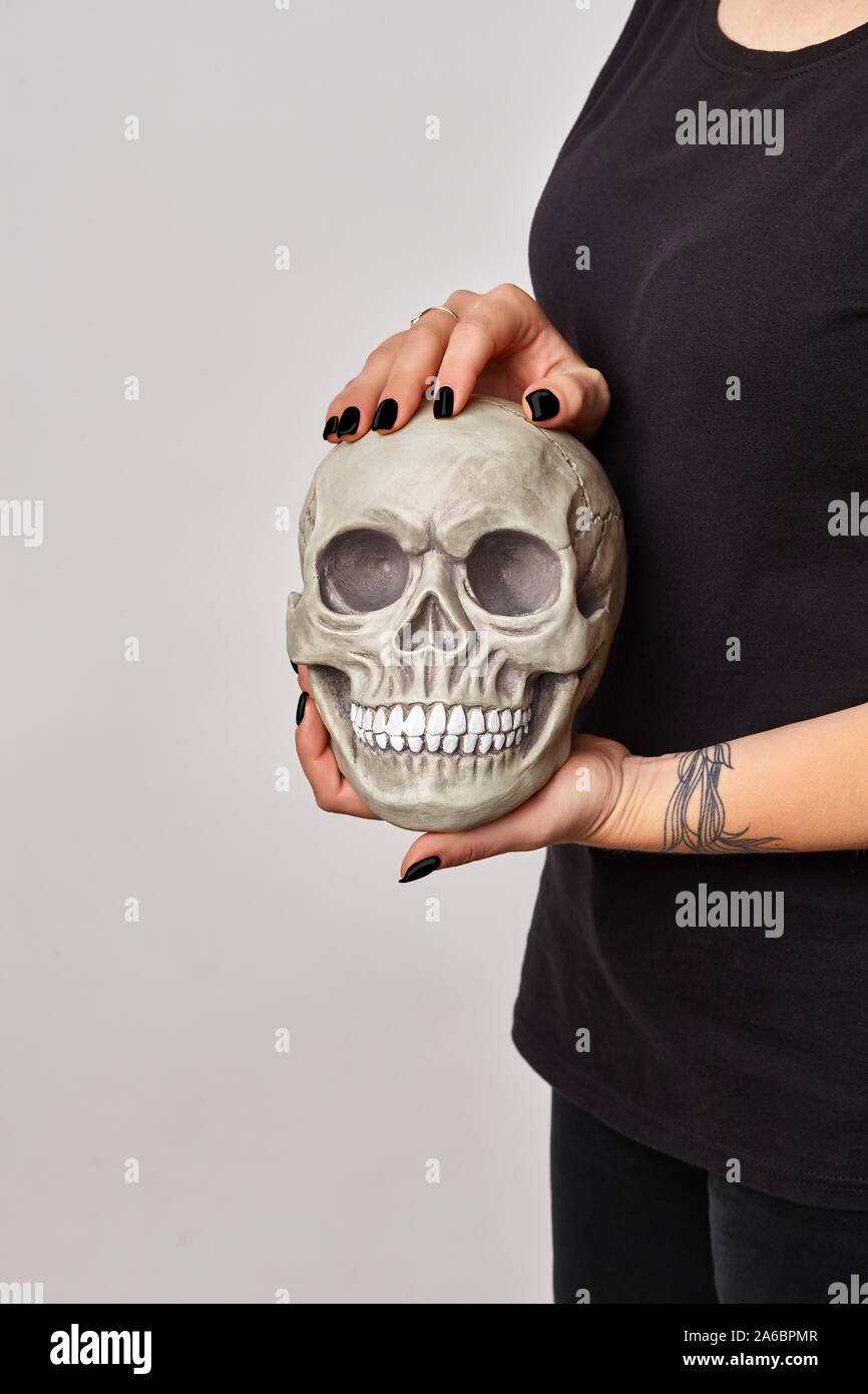 Tattooed hand of a female in a black watch and clothes is holding a realistic model of a human skull with teeth isolated on white. Medical science or Stock Photo