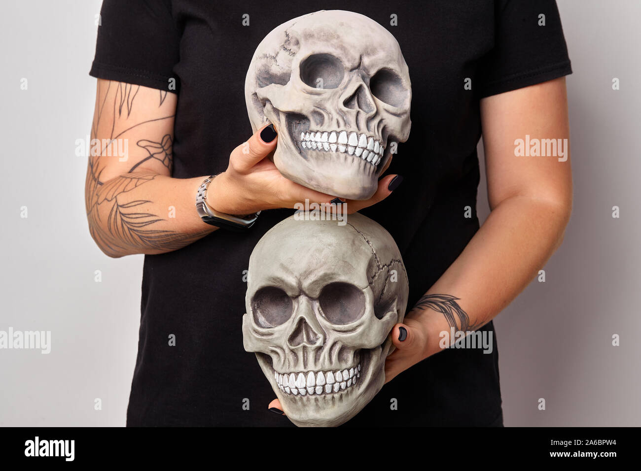 Tattooed hands of a woman in a black watch and clothes are holding a realistic model of two human craniums with teeth, isolated on white. Medical scie Stock Photo