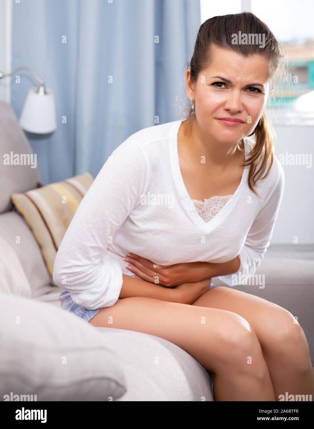 Young brunette suffering from abdominal pain, sitting on couch and holding her stomach with hands Stock Photo