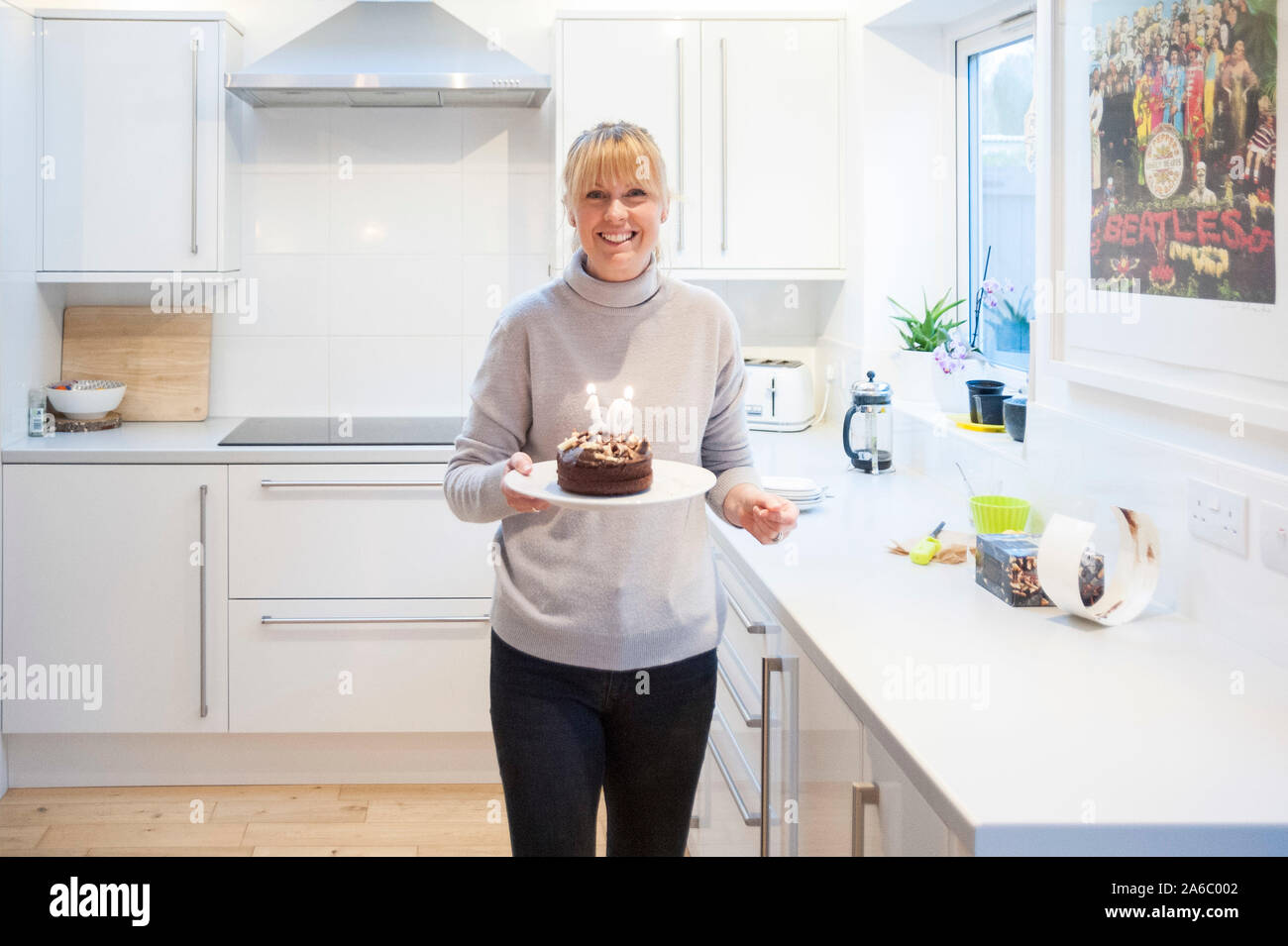 A mother stands in the kitchen with a birthday cake and the candles lit Stock Photo