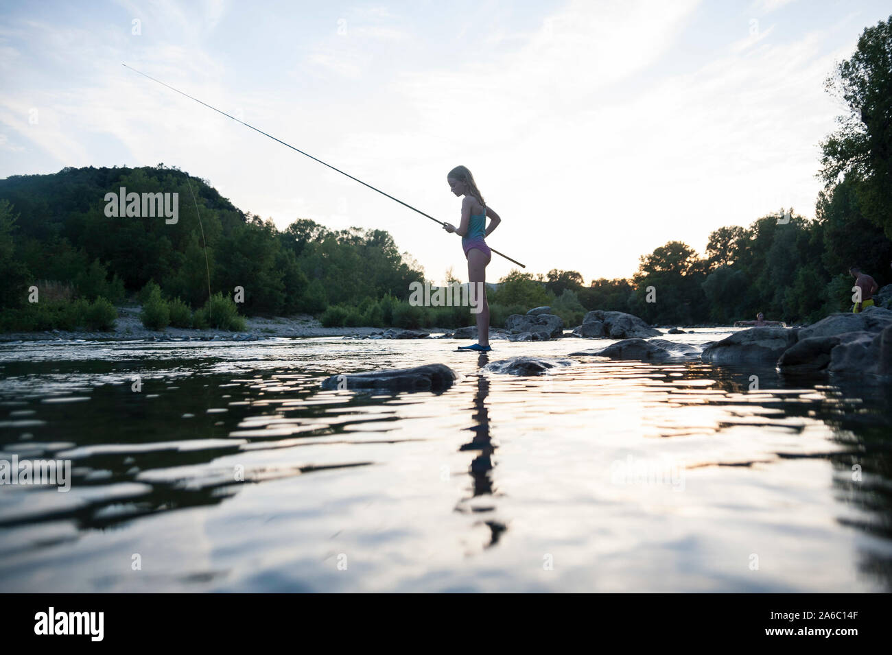 A kid starts ands at the waters edge at sunset fishing with a rod. Stock Photo