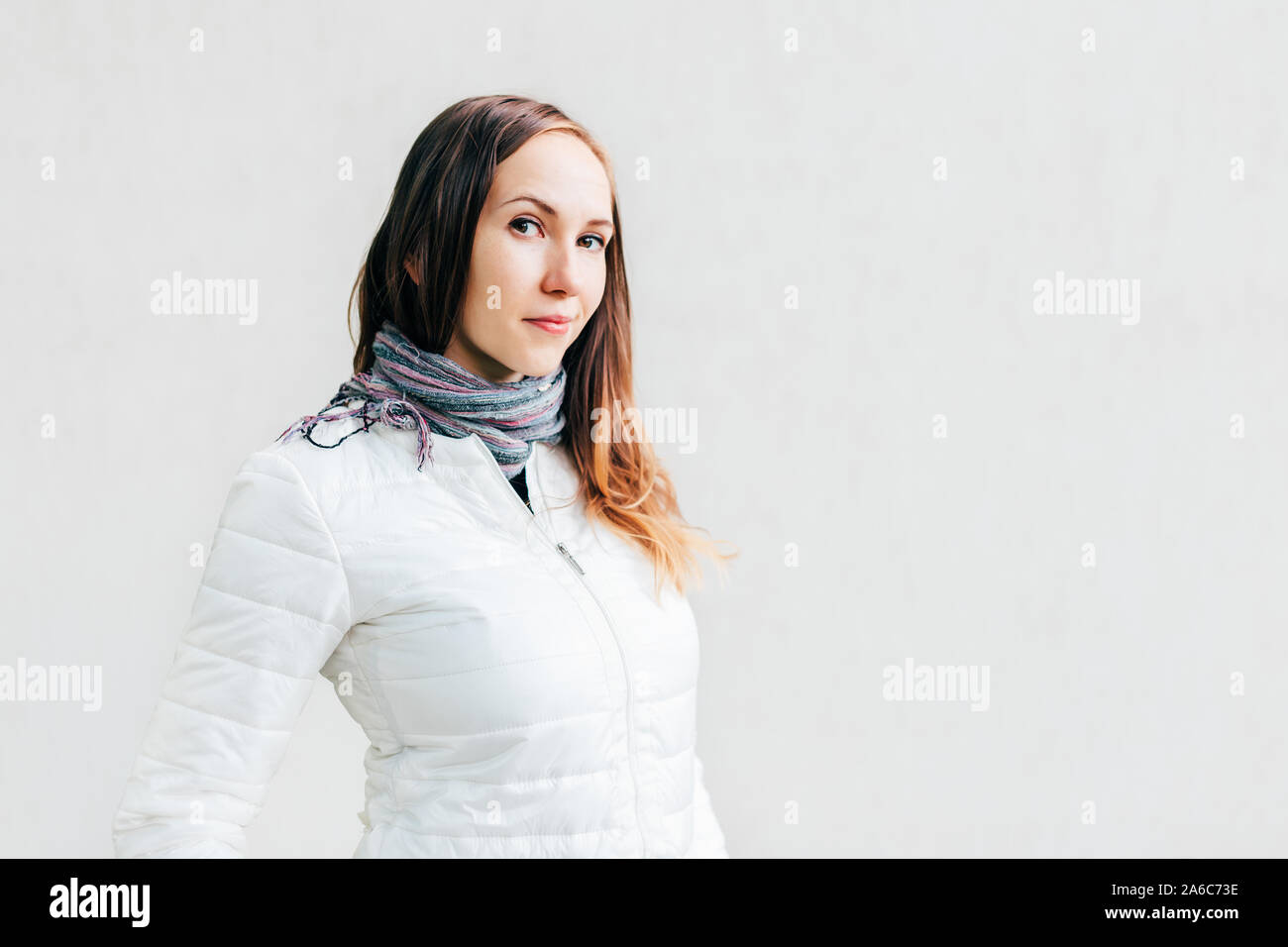 Young Caucasian woman in white jacket with long brunette hair looks into the lens with little smile on background solid monochrome wall. Stock Photo