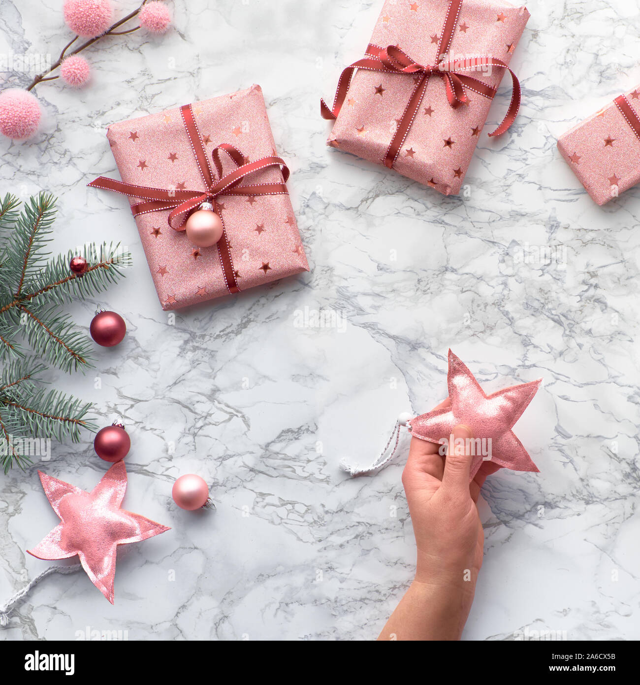 Christmas flat lay on marble table, square composition. Hand holding decorative Xmas star. Winter decorations: fir twigs and pink trinkets. Stock Photo