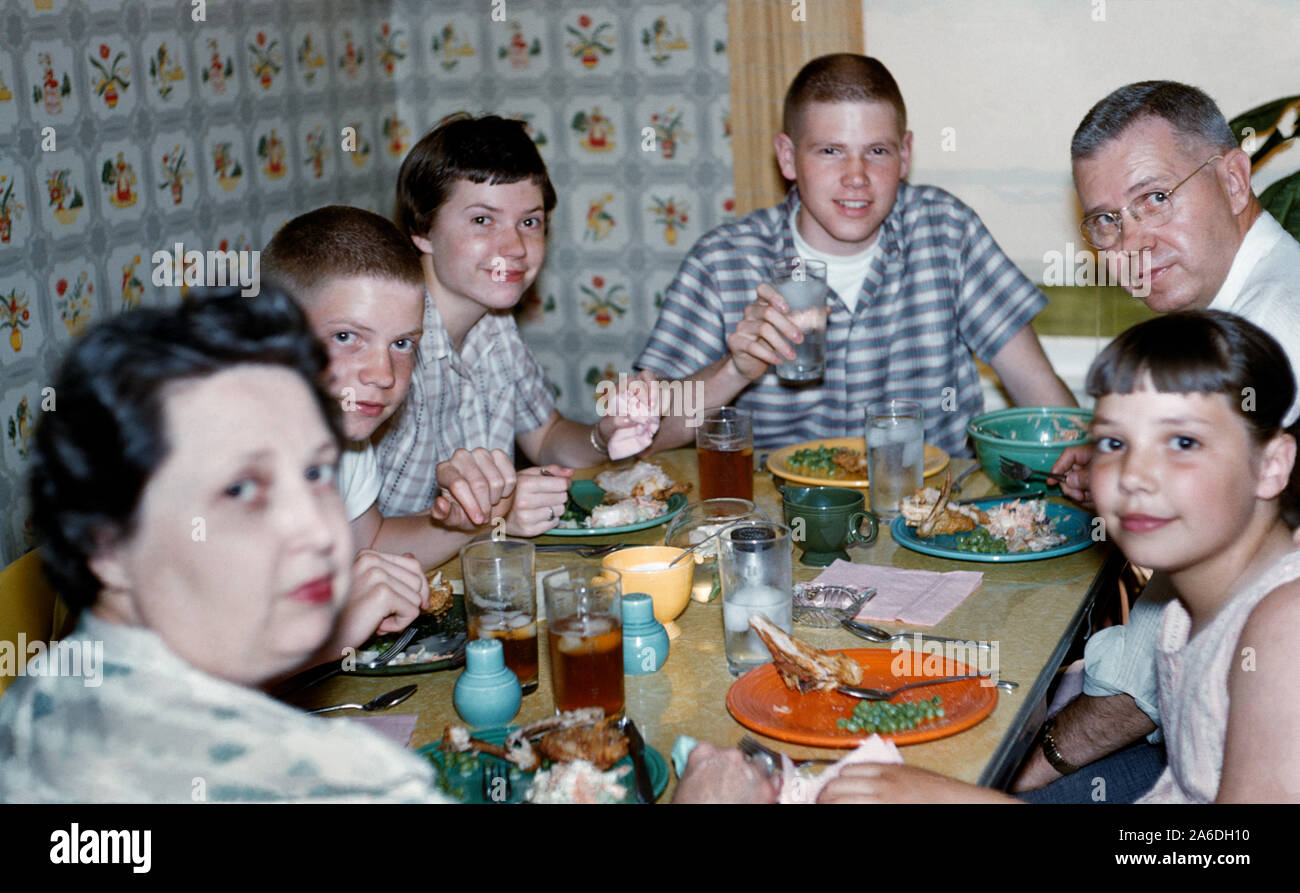 A 1950s Midwestern American family of six gather closely together for dinner around a small Formica-topped table to eat fried chicken, mashed potatoes and green peas served on multicolored Fiesta dinnerware with glasses of iced tea and water. Also note cigarette ashtray on the table, short hair styles, and patterned wallpaper typically in fashion during that mid-20th Century era. Historical photo. Stock Photo