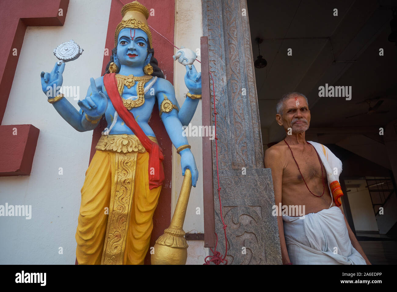 A priest stands in the entrance of Sri Venkataramana Temple in Car Street, Mangalore (Mangaluru), Karnataka, India, next to a statue of god Vishnu Stock Photo