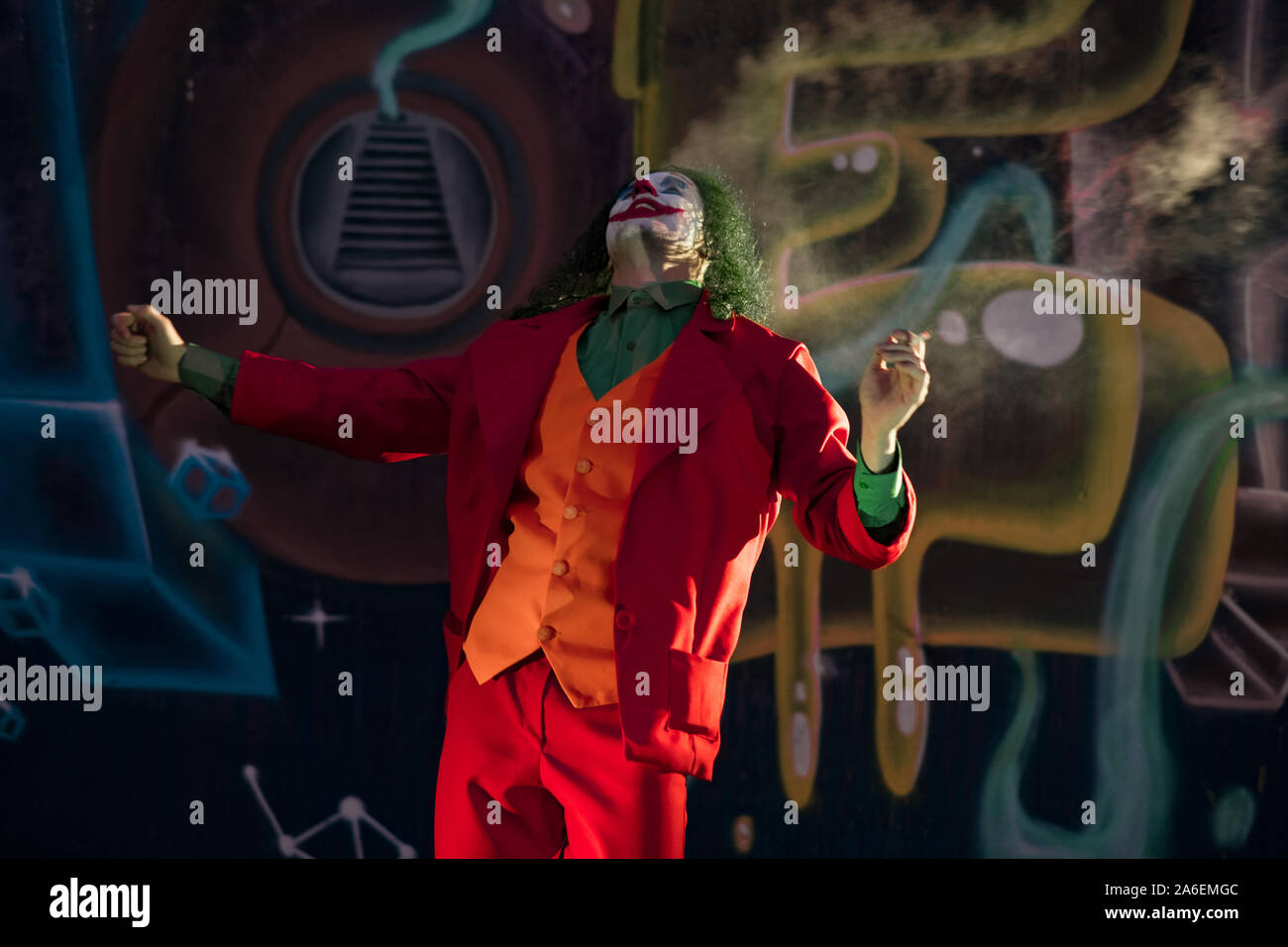 Cosplayer in the image of a crazy clown smokes a cigarette against  background of wall with graffiti Stock Photo - Alamy