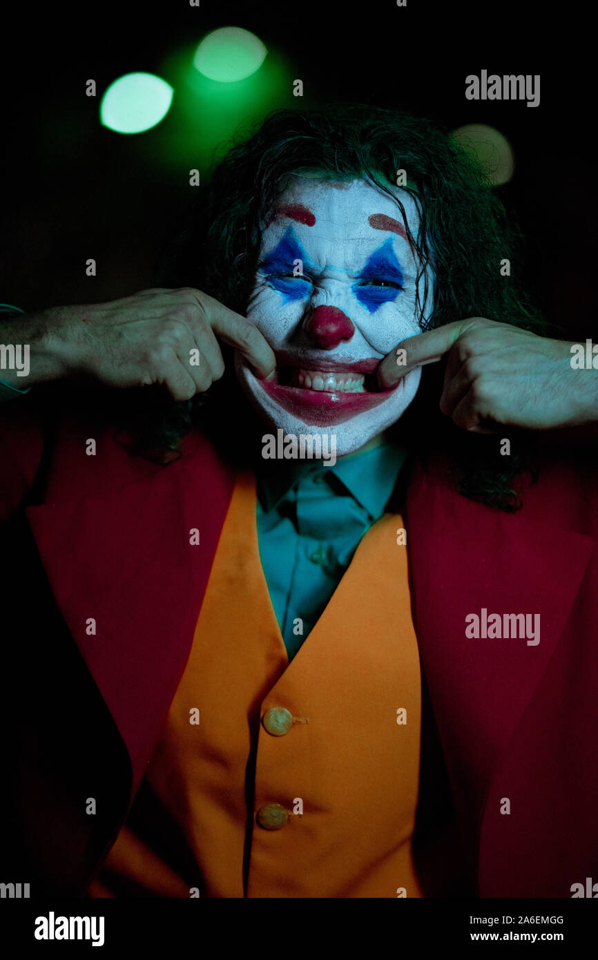 Cosplayer in the image of a crazy clown grimaces and smiles at nigth city. Stock Photo