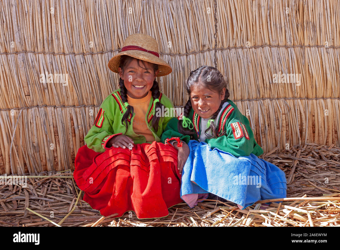 A portrait of two young girls sitting in front of a straw hut on a floating Uro Island in Lake Titicaca, Puno, Peru. Stock Photo